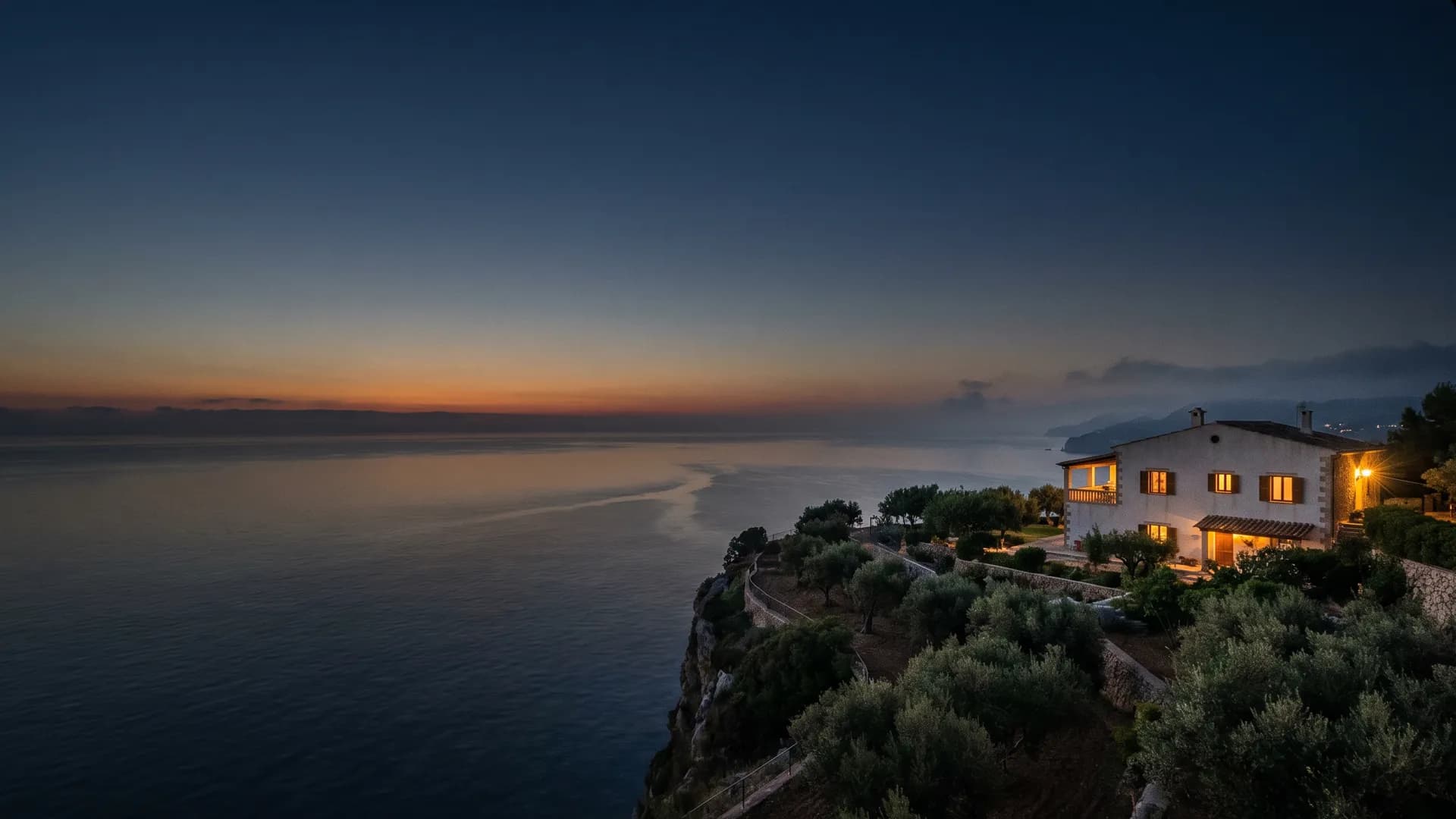 First light over a calm Mediterranean sea; a coastal home on the headland, its windows warmly lit